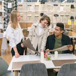 Three young adults discuss table options in a stylish furniture store, symbolizing teamwork and modern interior design.